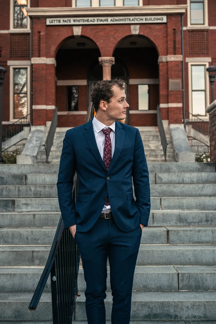 Daniel i kostym och examensstola framför en universitetsbyggnad.